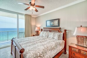 Une chambre avec un grand lit, un ventilateur de plafond, une table de chevet avec une lampe, un tableau encadré au mur et une vue sur l’océan par une grande fenêtre.