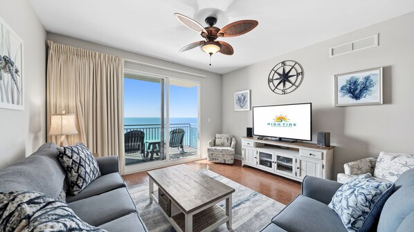 A modern living room with a large TV, a ceiling fan, and a view of the ocean through the sliding glass doors. - *Oct 28 - Nov 5th open! * Beautiful condo! (Panama City Beach)
