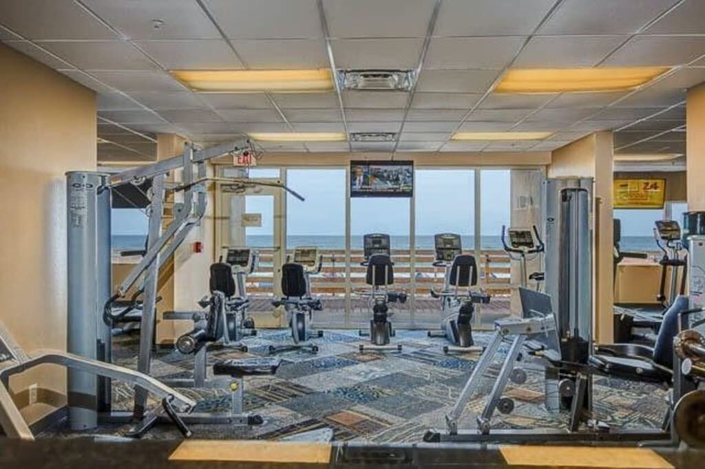 A gym with various exercise machines, a television mounted on the wall, and a view of the ocean through large windows.