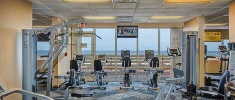A gym with various exercise machines, a television mounted on the wall, and a view of the ocean through large windows.