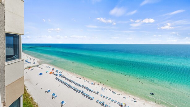 Ein Strand mit klarem türkisblauem Wasser, ein Pier in der Ferne sowie Strandstühle und Sonnenschirme.
