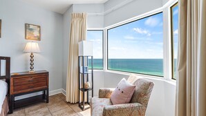Ein Zimmer mit großem Fenster und Meerblick, ein gemustertes Sessel, ein Nachttisch mit Lampe und ein Regal mit Dekoration.