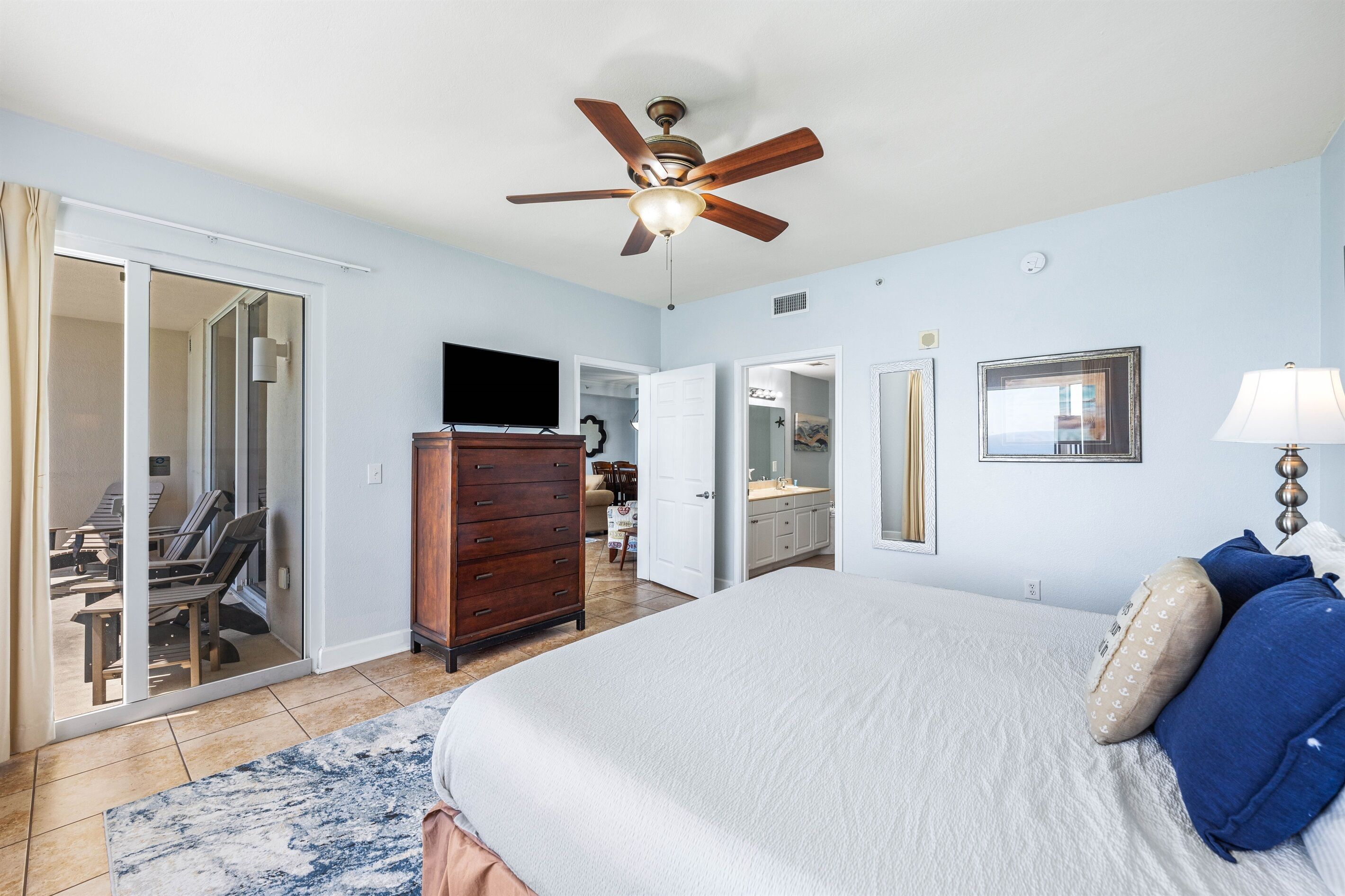 A bedroom with a bed, a dresser, a ceiling fan, a television, and a bathroom visible through a glass door.