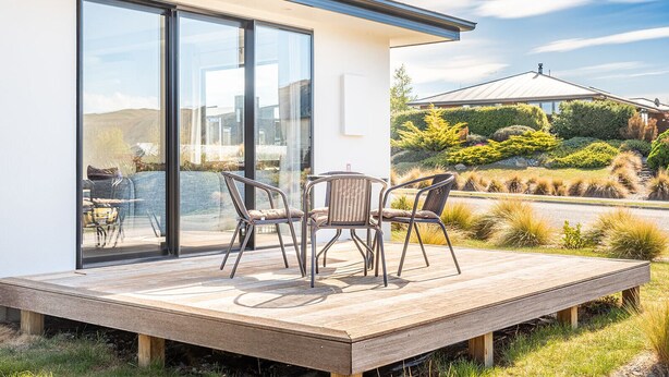 A modern house with a wooden deck, two chairs, and a glass door leading to a garden.