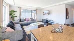 A modern living room with a grey sofa, a wooden dining table, and a large window with curtains.