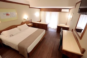 A hotel room with a bed, bedside tables, a desk, a chair, a mirror, and a window with curtains.
