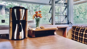 A kitchenette with a stainless steel coffee maker, a wooden cutting board with pastries, and a vase with flowers by a window. - AGS Trailer House - Campground (Hakuba)