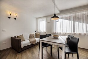 A modern living room with a sofa, dining table, chairs, pendant lighting, and a large window with sheer curtains.