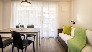 Ein Zimmer mit Esstisch, Stühlen, einer Couch, einer grünen Decke und einem Fenster mit durchsichtigen Vorhängen.