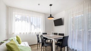 A modern room with a dining table, chairs, a sofa with green cushions, a flat-screen TV, and sheer curtains. - Apartment "Set Residenz Bun Sté" with Mountain View, Sauna & Balcony (San Vigilio di Marebbe)