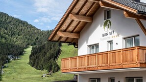 A hotel with a wooden balcony, a mountain view, and a logo with a tree and a person. - Apartment "Sis Residenz Bun Sté" with Mountain View and Sauna (San Vigilio di Marebbe)