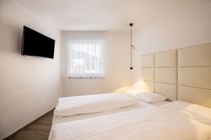 A hotel room with two beds, a flat-screen TV mounted on the wall, and a window with sheer curtains.