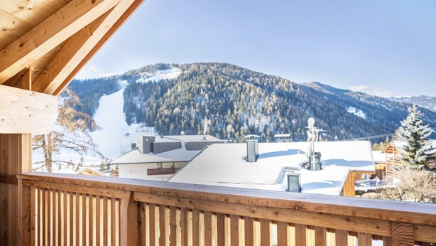 A wooden balcony with a view of a snow-covered mountain and buildings. - Apartment "Sis Residenz Bun Sté" with Mountain View and Sauna (San Vigilio di Marebbe)