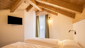 A bedroom with two beds, a flat-screen TV, a window with curtains, and a wooden ceiling. - Apartment "Sis Residenz Bun Sté" with Mountain View and Sauna (San Vigilio di Marebbe)