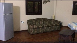 A room with a wooden floor, a grey refrigerator, a camouflage sofa, a small wooden table, and a window with a decorative wall hanging. - Casa in Masca Omoa (Omoa)
