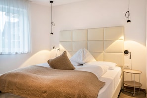 Un lit bien fait, avec une tête de lit, une table de chevet sur laquelle se trouve une lampe, et une fenêtre ornée de rideaux légers.