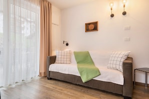 Une pièce comprenant un canapé, un plaid vert, un tableau encadré en bois, des lampes fixées au mur et une fenêtre ornée de rideaux légers.