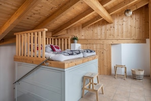 Une chambre à coucher aménagée dans un espace de type « loft », avec un plafond en bois, un lit avec des oreillers, une petite table, un tabouret et un panier.
