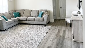 Un soggiorno con un divano angolare grigio, un tappeto decorato, una ventola a soffitto e un pavimento in legno.