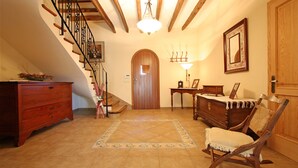 Una escalera con barandillas de madera y un candelabro, una puerta de madera, una mecedora, un aparador de madera y una mesita con una lámpara.