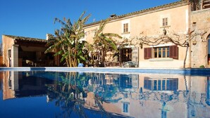 Una piscina refleja un edificio con techo y contraventanas de azulejos, rodeado de vegetación.