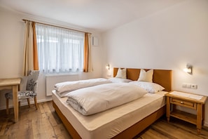 A bedroom with a double bed, a desk, a chair, a nightstand, and a window with curtains.