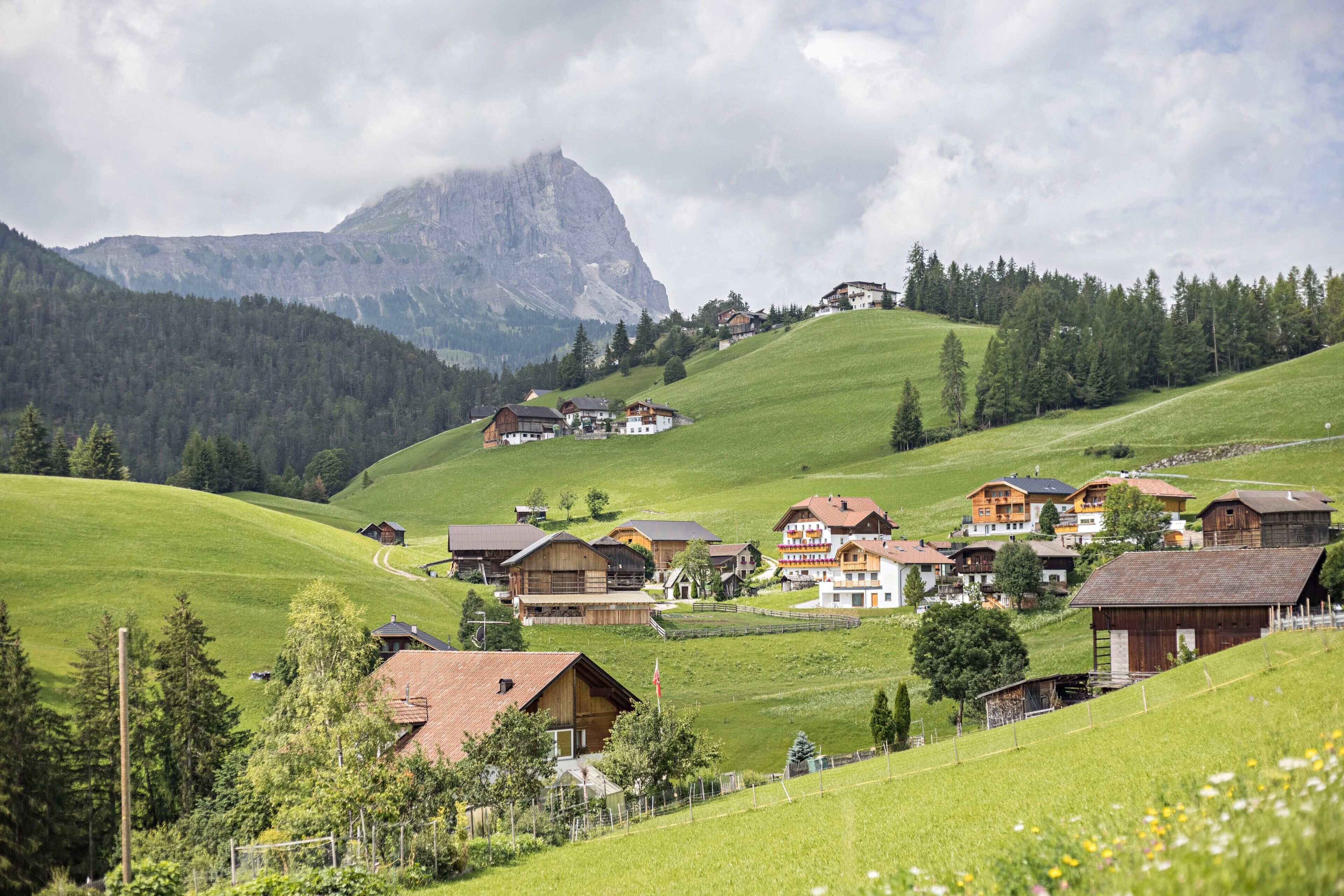 Un villaggio rurale con case in legno tradizionali, una montagna imponente sullo sfondo e cielo sereno.