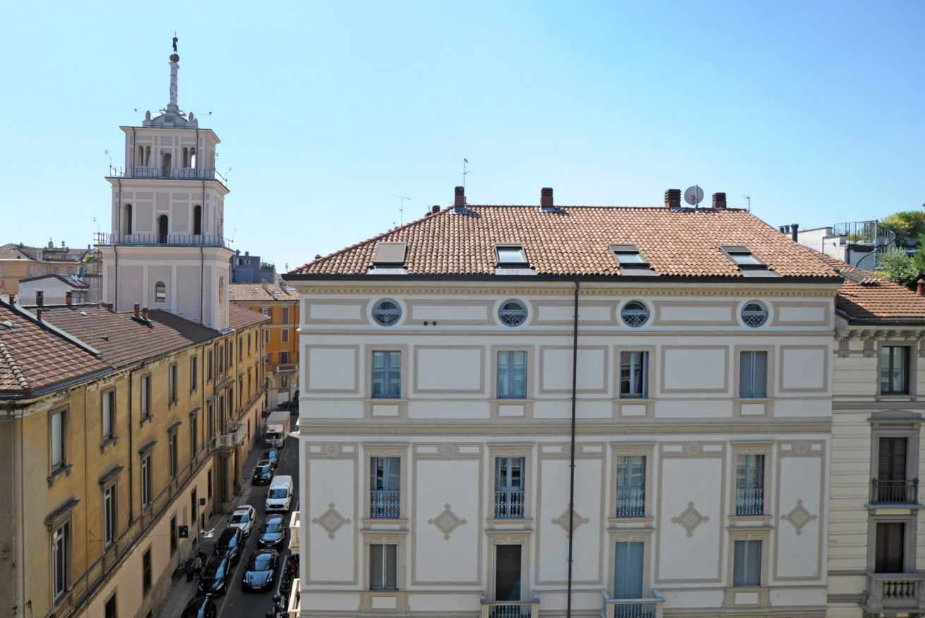 A multi-story building with a tiled roof, a bell tower, and a street with parked cars.