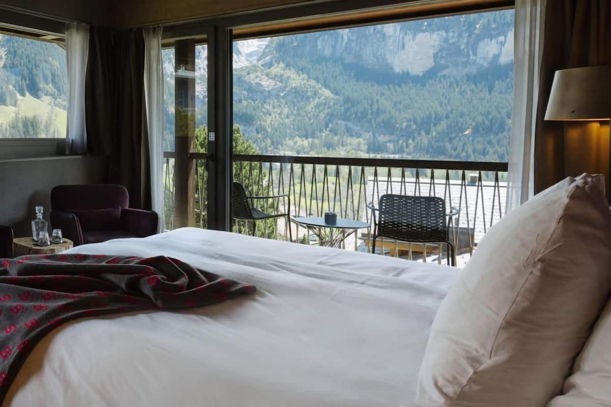 Habitación de hotel con una cama grande, un balcón con vista a la montaña y una mesa con sillas.