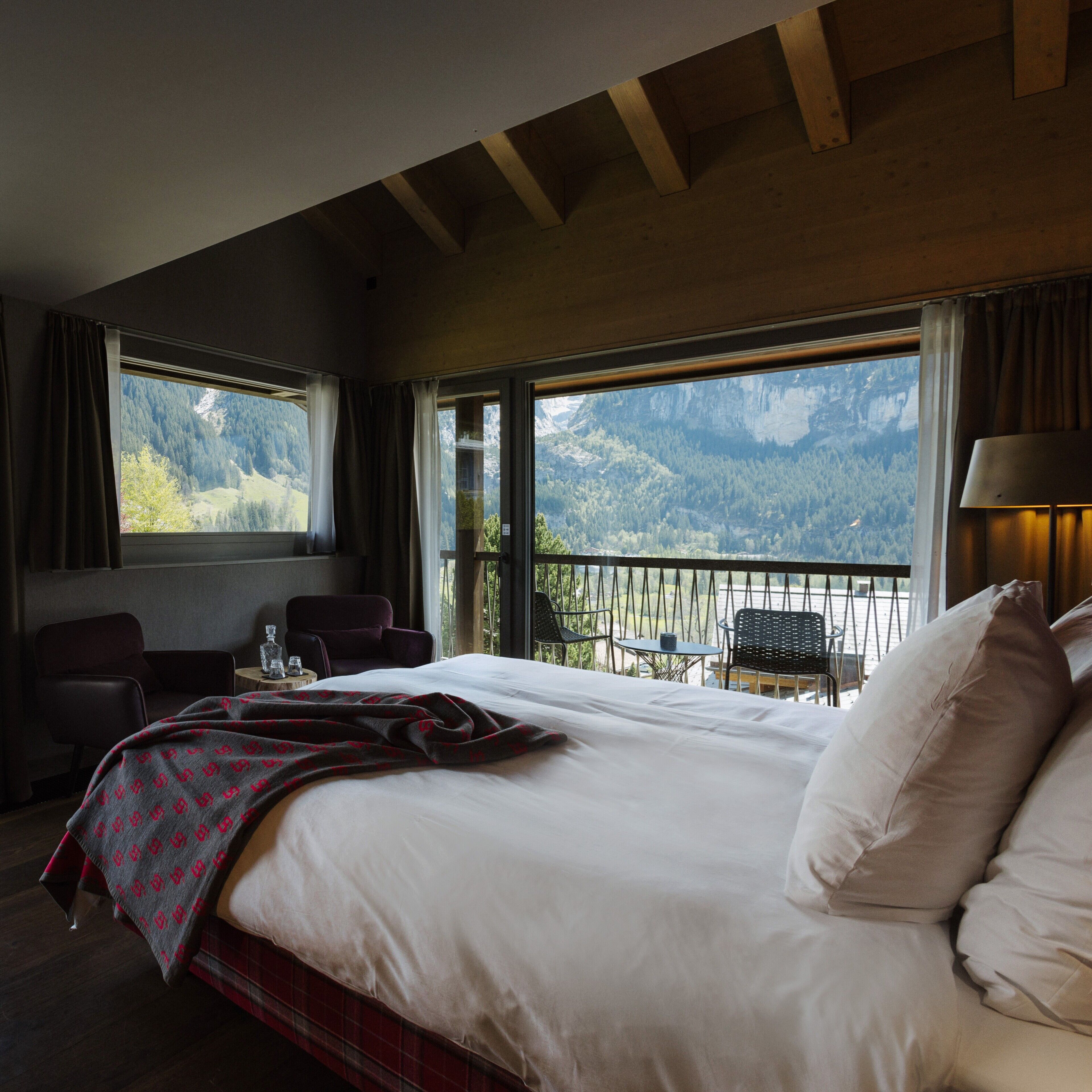 A hotel room with a large bed, a view of mountains, a balcony with chairs, and a table with a glass.