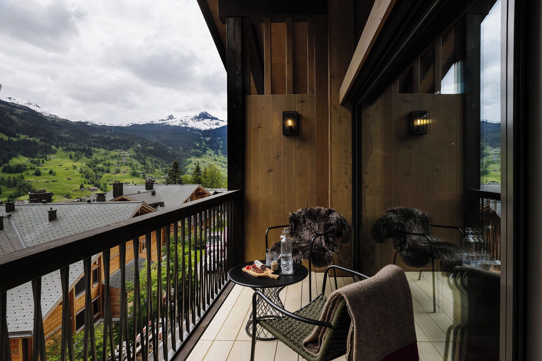A balcony with a view of a mountainous landscape, a table with a bottle, glasses, and a plate of food, and a chair with a blanket.