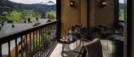 A balcony with a view of a mountainous landscape, a table with a bottle, glasses, and a plate of food, and a chair with a blanket.