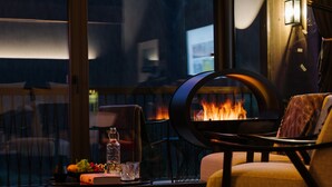 A cozy room with a fireplace, a comfortable chair, a small table with a fruit bowl, a bottle, and a glass.