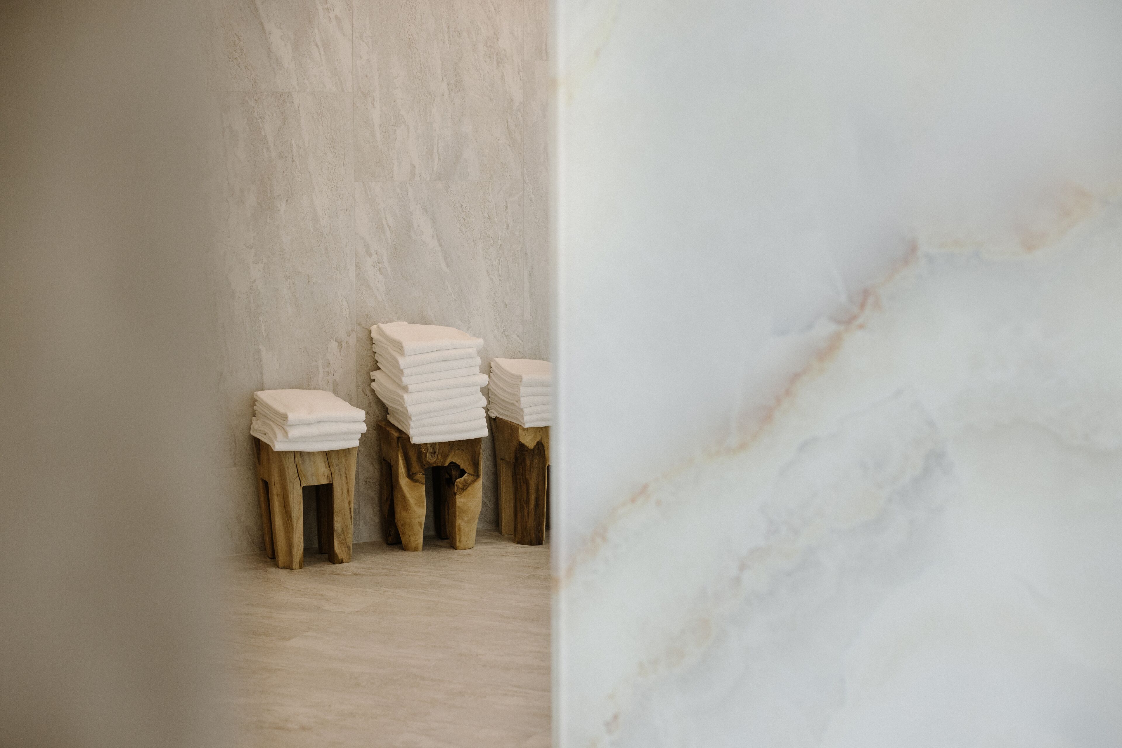 Rincón de una habitación con pared de mármol y un taburete de madera con toallas blancas dobladas.
