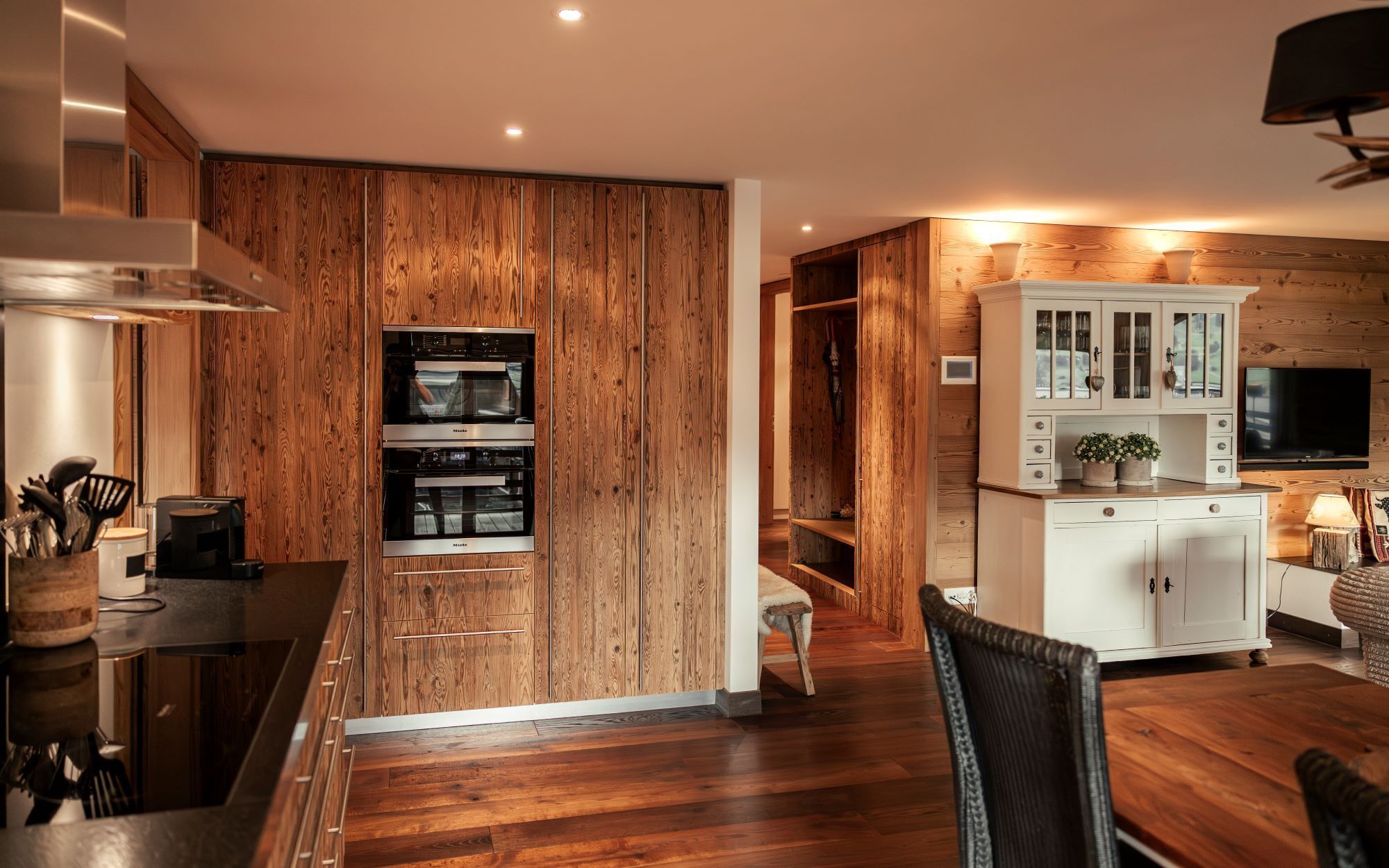 A modern kitchen with wooden paneling, built-in ovens, and a white cabinet with glass doors.