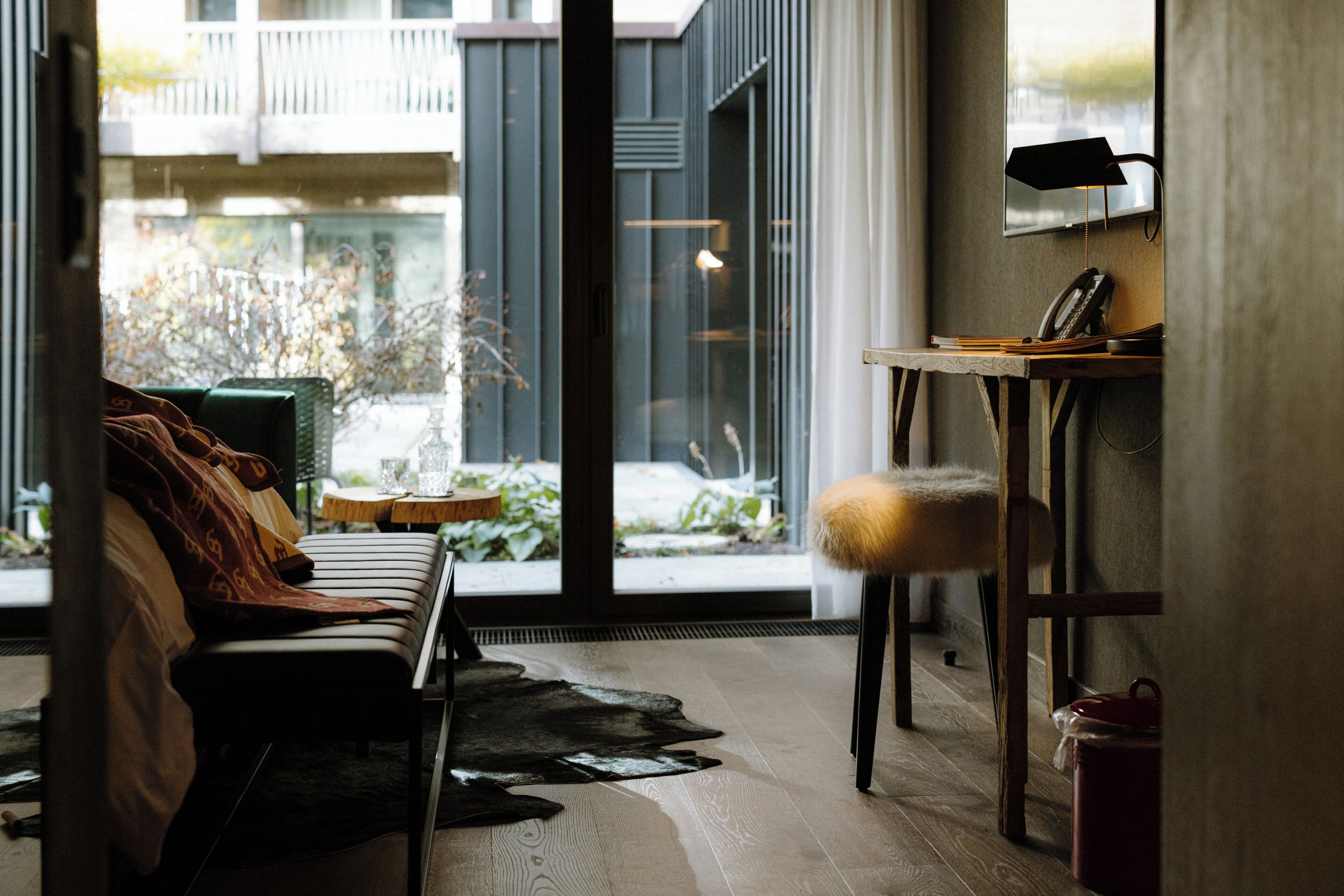 A modern living room with a sofa, a small table, and a chair. There is a view of the outside through large windows.