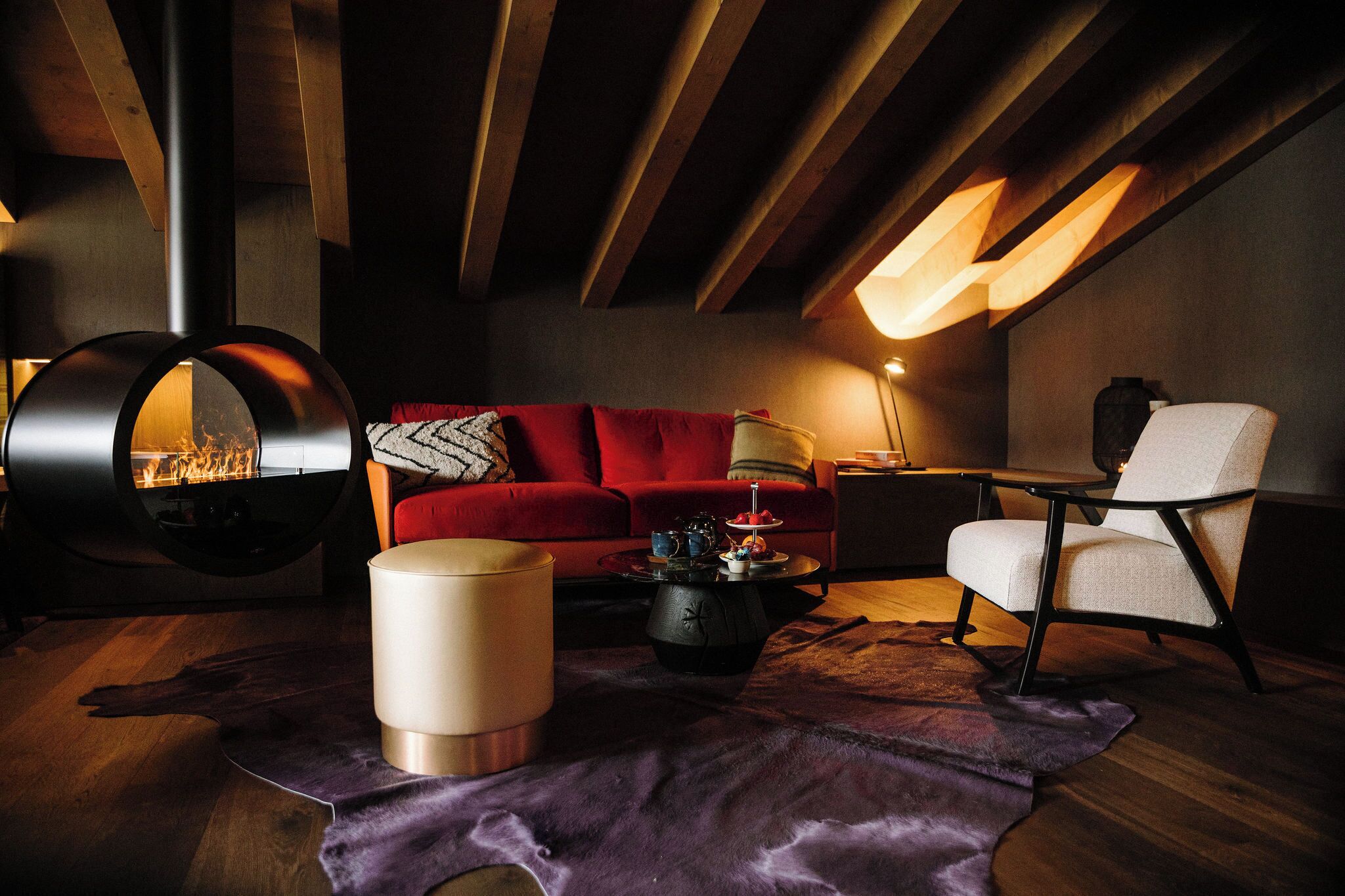 Un salon chaleureux avec un canapé rouge, une cheminée ronde, un fauteuil blanc et un tapis en cuir.