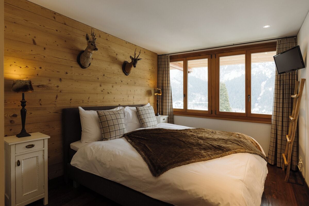 A bedroom with a bed, wooden wall paneling, mounted animal heads, a nightstand, a television, and a window with a view of mountains.
