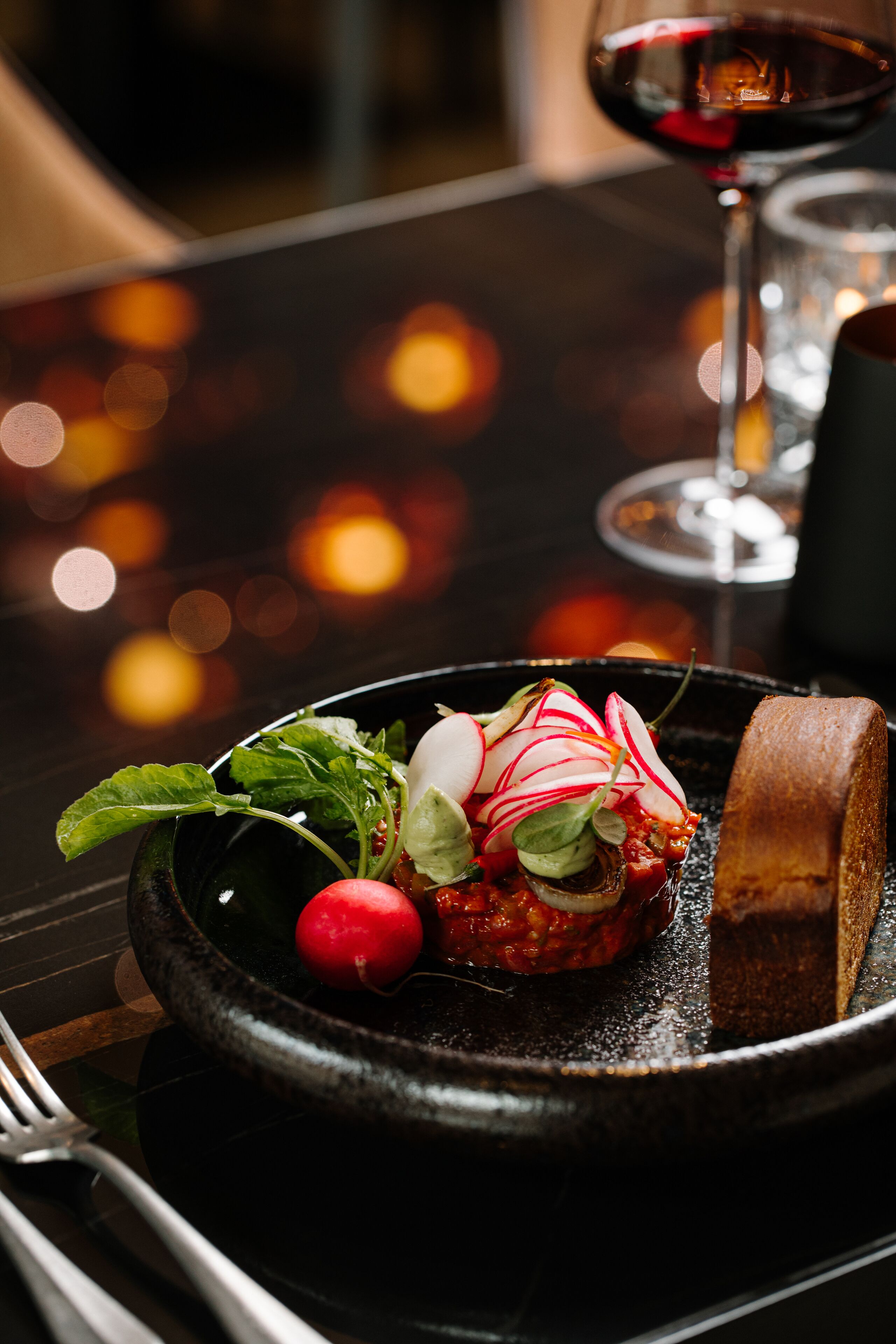 A dining table with a black plate containing a dish with radishes, greens, and a slice of bread, and a glass of red wine in the background.