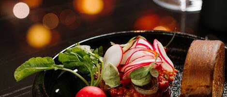A dining table with a black plate containing a dish with radishes, greens, and a slice of bread, and a glass of red wine in the background.
