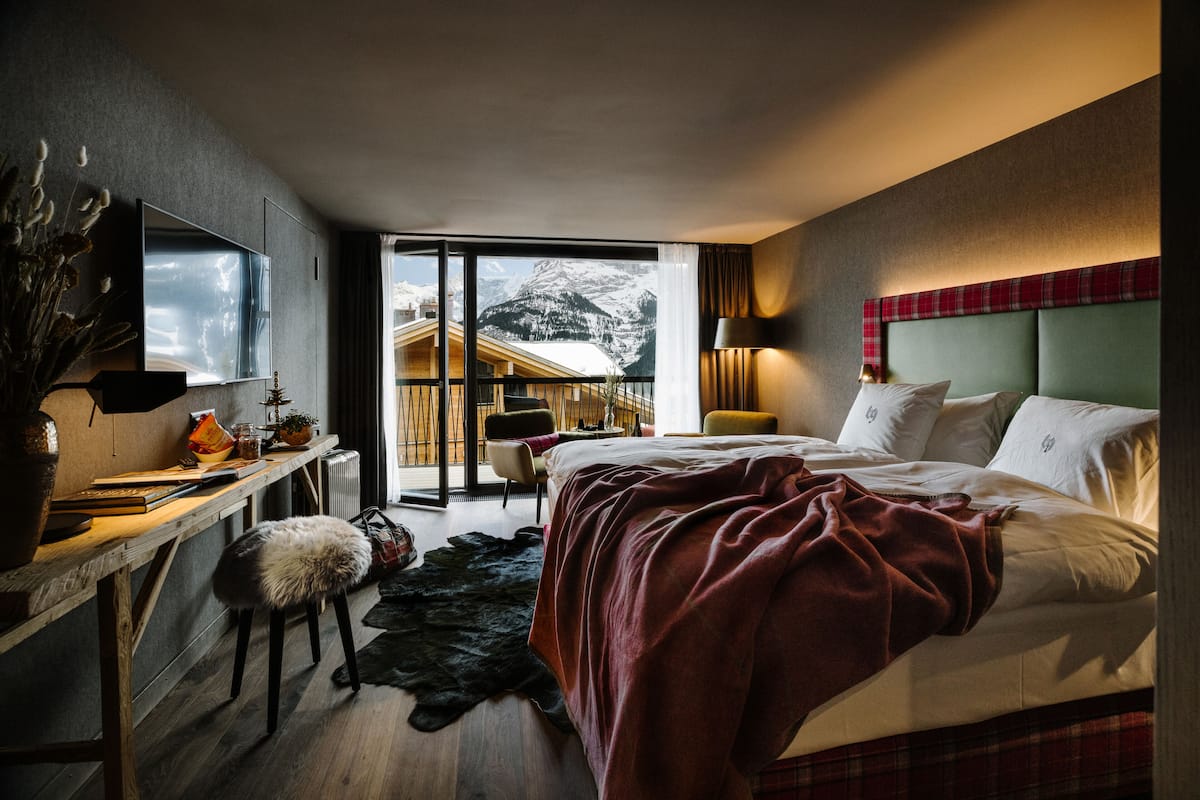 Una habitación de hotel moderna con una cama grande, un escritorio con una silla, un televisor montado en la pared y vistas a la montaña a través de una puerta corredera.