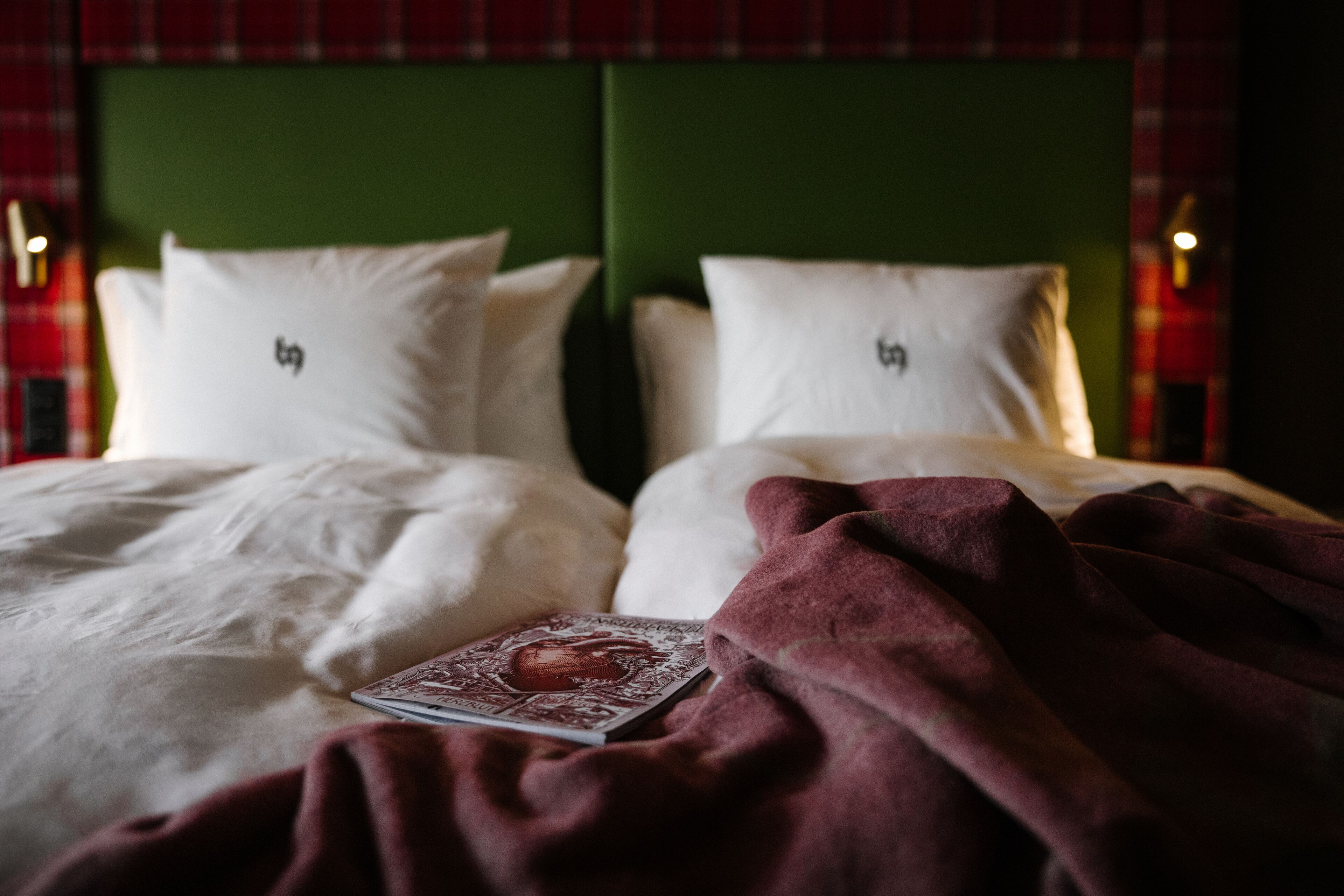 A bed with white bedding and a red blanket, a book, and pillows with a logo.