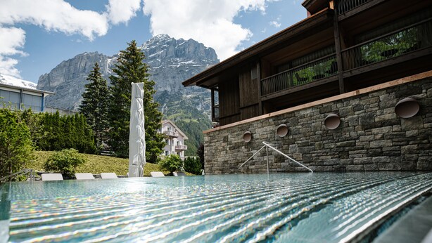 A pool with a mountain backdrop, stone walls, and a wooden building with balconies.