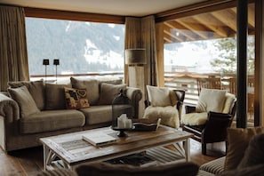 A cozy living room with a sofa, armchair, and coffee table, featuring a view of mountains through large windows.