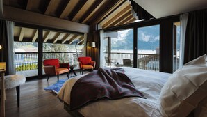 A bedroom with a large bed, wooden ceiling beams, a balcony with chairs, and a view of mountains.