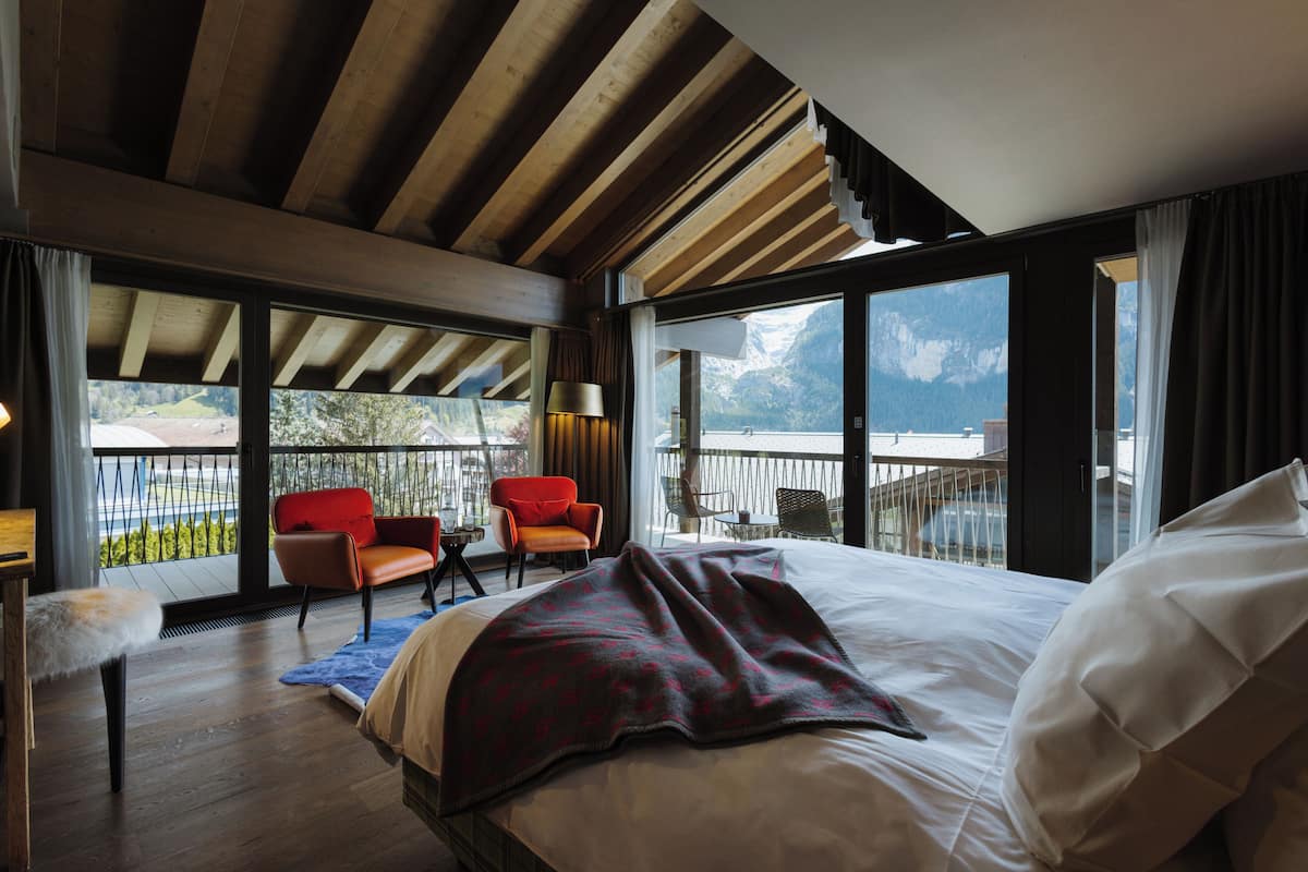 Un dormitorio con una cama grande, vigas de madera en el techo, un balcón con sillas y vistas a las montañas.