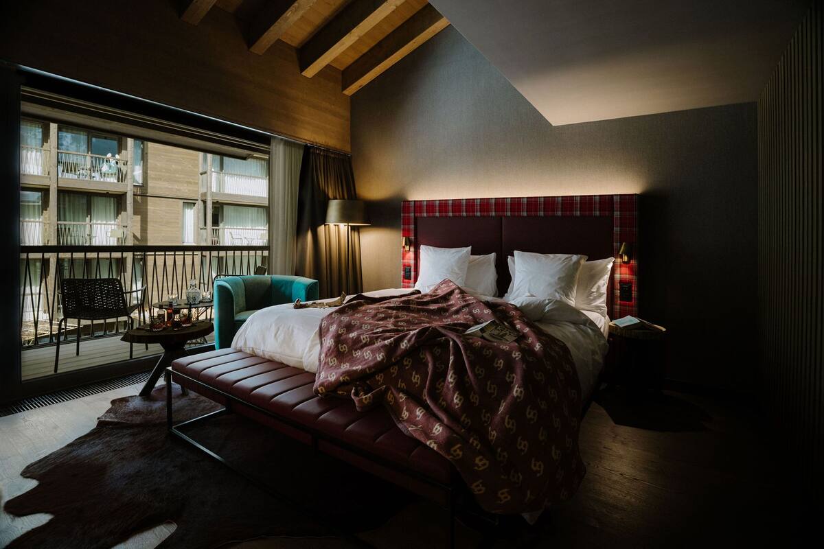 Una habitación de hotel moderna con una cama grande, un sillón de descanso, vista a un edificio con balcones y una ventana con cortinas.