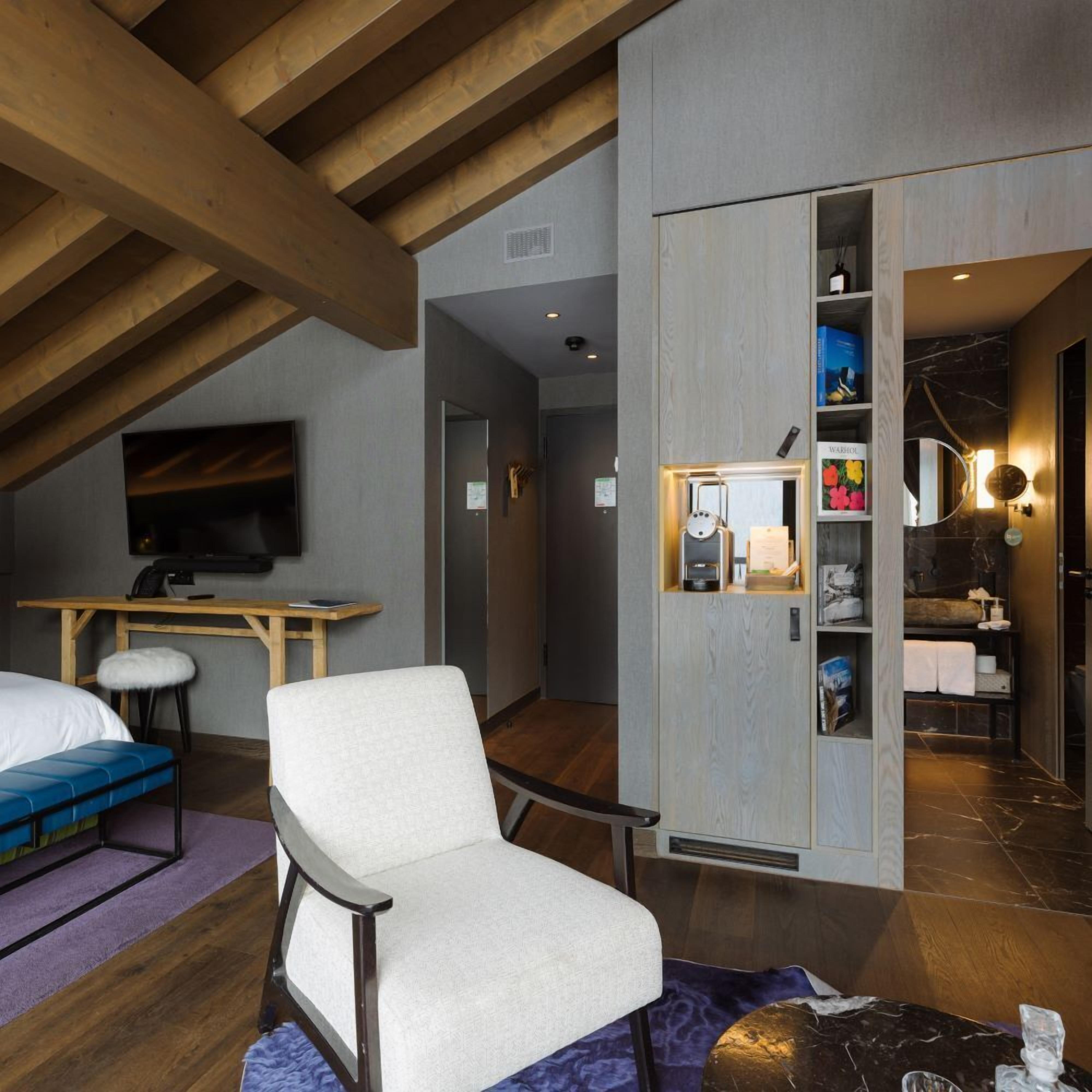 A modern hotel room with a loft-style ceiling, a large bed, a desk, and a seating area with a chair and a sofa.