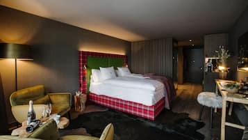 A modern hotel room with a large bed, a green upholstered chair, a small table with a bottle and glasses, and a wooden desk with a lamp.