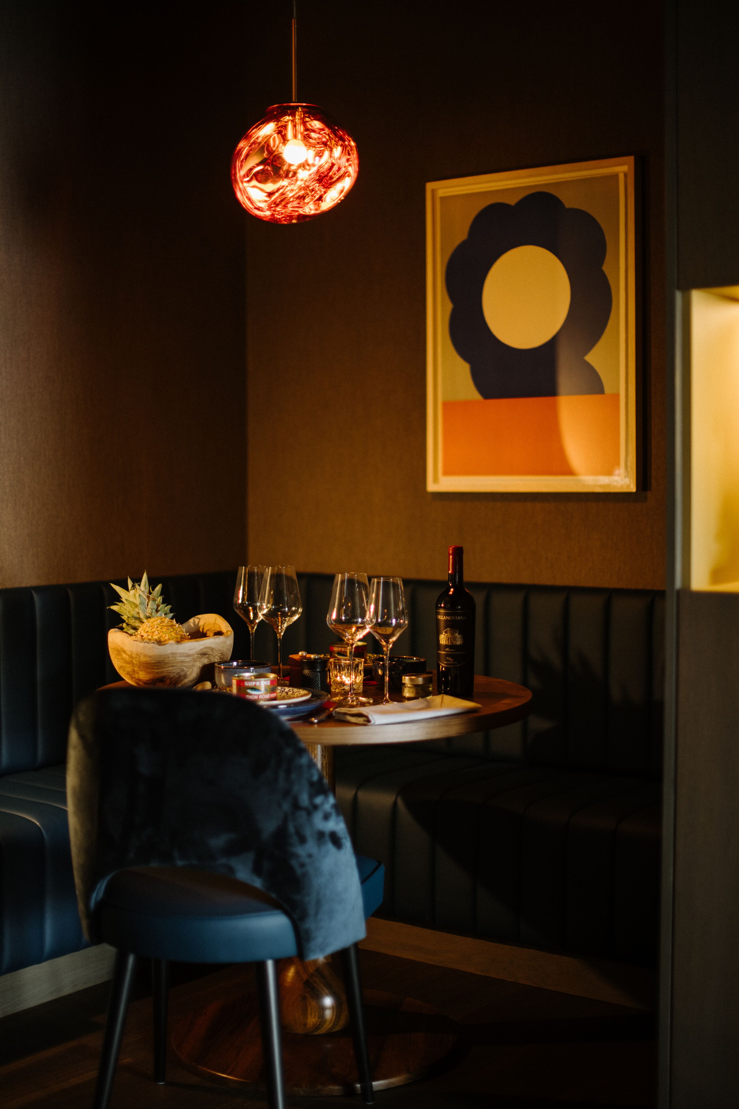 A dimly lit restaurant with a round table set for two, a bowl of fruit, wine glasses, and a bottle of wine.